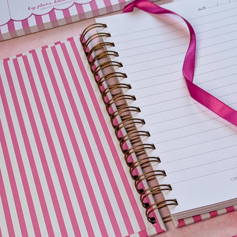 Pink and white striped notebook with a pink ribbon, pen, and decorative paper clips on a matching background.