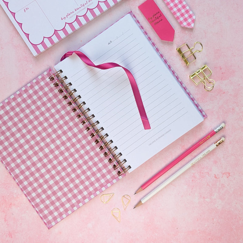 Open notebook with pink gingham cover and accessories on a pink background