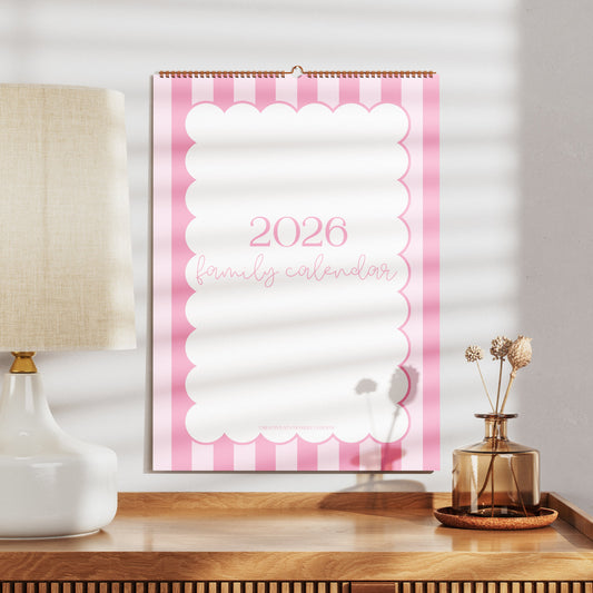 2026 wall calendar with pink and white design on a wooden surface with a lamp and decorative items.