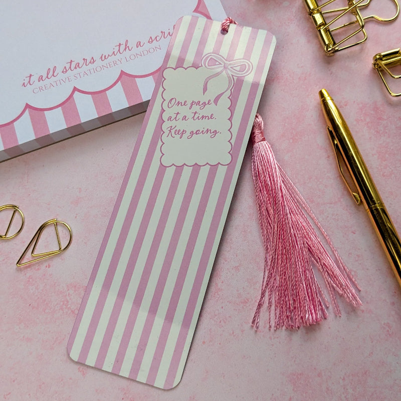 Striped bookmark with pink tassel on a pink background with stationery items.