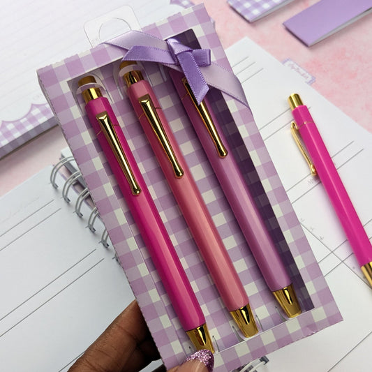 Set of pink pens with gold accents in a checkered box on a pink surface.