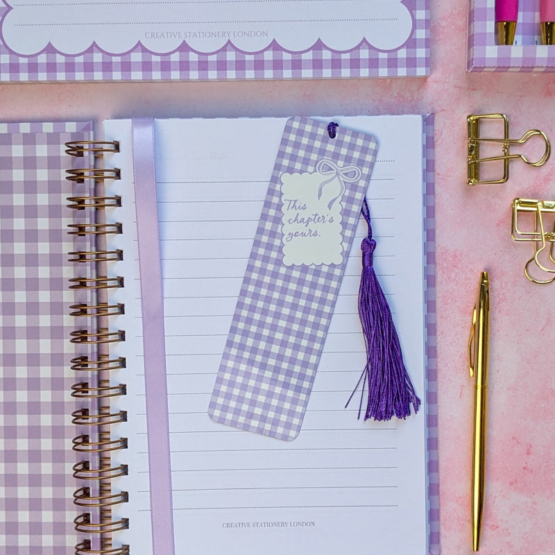 Notebook with a gingham bookmark, gold pen, and clips on a pink and purple checkered background.